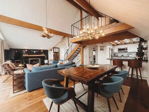 A spacious living area with a wooden dining table, blue sofas, and a staircase leading to an upper level. - Harlow House, w/ hot tub, lake view & lake access! (Lake Arrowhead)