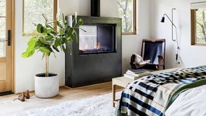 Ein gemütliches Schlafzimmer mit Kamin, großen Fenstern, einem Blumentopf und einem Bett mit gestreifter Decke.