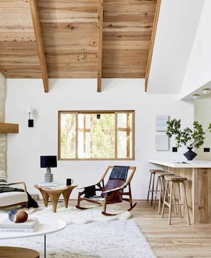 A spacious living room with a high ceiling, a large window, a wooden rocking chair, a bar area, and a cozy fireplace. - The Mountain House, by designer Emily Henderson (Lake Arrowhead)