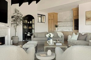 A living room with a fireplace, sofas, a coffee table, and a kitchen area in the background. - Pinnacle Point Lake House (Lake Arrowhead)