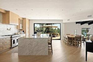 A modern kitchen with a marble island, wooden cabinets, and a dining area with a large window overlooking a garden. - Pinnacle Point Lake House (Lake Arrowhead)