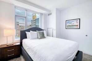 Ein modernes Hotelzimmer mit einem großen Bett, einem Nachttisch, einer Lampe, einem Fenster mit Blick auf die Stadt und einem gerahmten Bild an der Wand.
