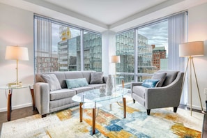 Ein modernes Wohnzimmer mit einem Glastisch, grauen Sofas und Blick auf eine Stadtlandschaft durch große Fenster.