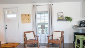 A room with a door, two wooden chairs with cushions, a patterned rug, a window with curtains, a coffee machine, and a shelf with a plant. - Peaceful Ozark Escape Near the Artesian Well, 2BR/2BA Retreat (Marquand)