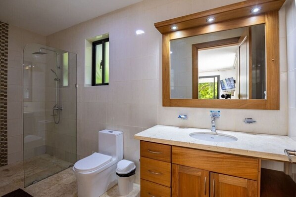 A modern bathroom with a glass shower enclosure, a toilet, a sink with a wooden vanity, and a mirror with a wooden frame. - Casa Coco Pace - Casa Lina 772 (Sosúa)