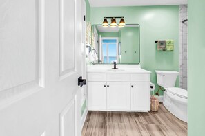 A bathroom with white cabinets, a green wall, a mirror with lights, a toilet, and a wooden floor. - 🦞Gulf Front | Beach Service | Pool | 2BR Ocean Villa 2101🌅 (Panama City Beach)