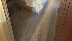Un dormitorio con cabecera de madera, cama con ropa blanca, radiador, ventana con cortinas y ventilador de techo.