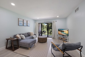 Ein modernes Wohnzimmer mit grauem Sofa, Hocker aus Weidengeflecht, grauem Sessel, Flachbildfernseher und großem Fenster mit Blick auf Grünflächen.