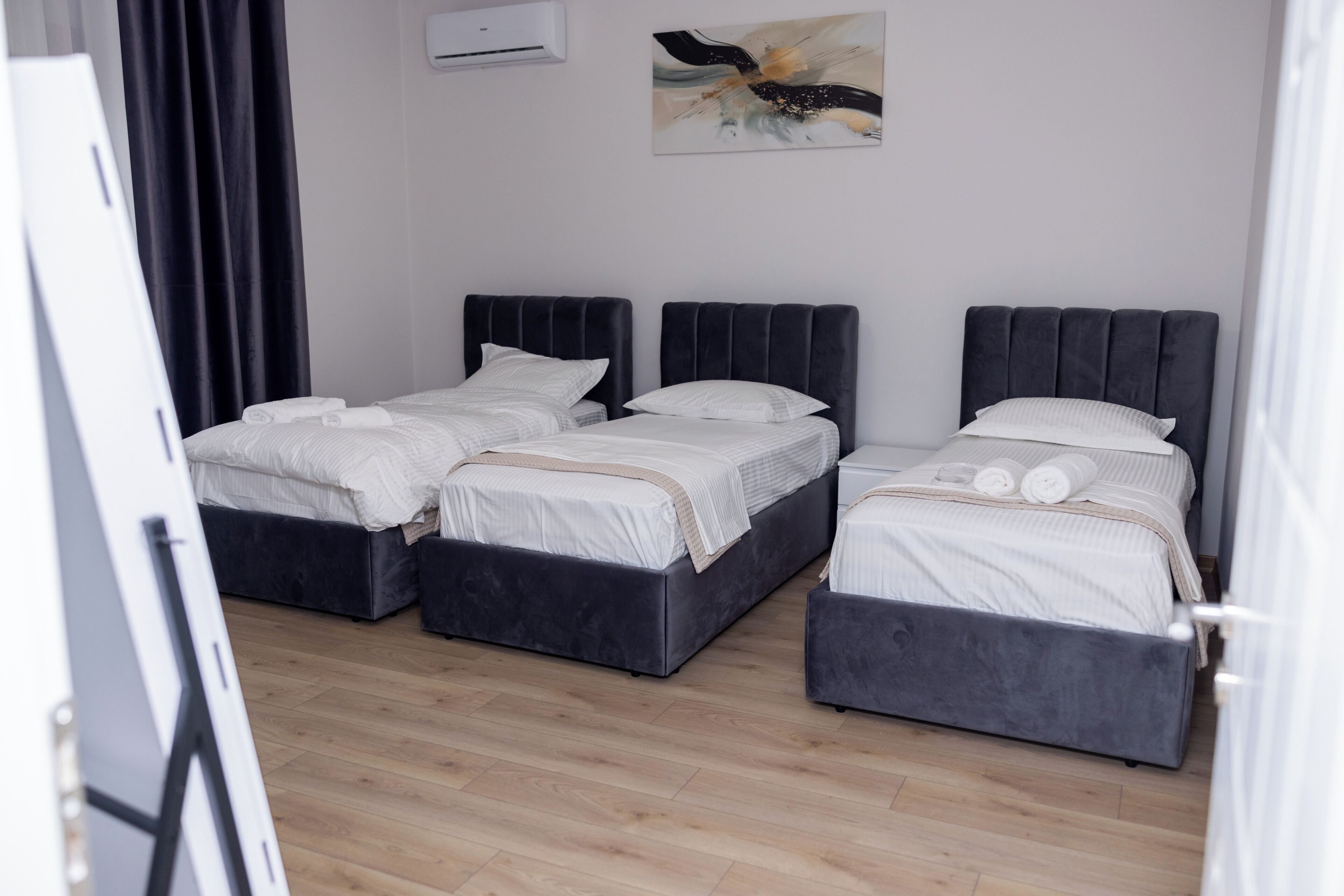 A hotel room with three beds, a wooden floor, a painting on the wall, and an air conditioning unit.