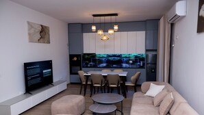 A modern living room with a sofa, ottoman, TV, dining table, chairs, and a kitchen area with a waterfall feature. - Modern 110 sqm apartment near Tirana and airport (Valias)