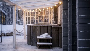 Eine hölzerne Außen-Sauna mit einer Sitzbank, einer Glastür mit herzförmigen Verzierungen und Lichterketten.