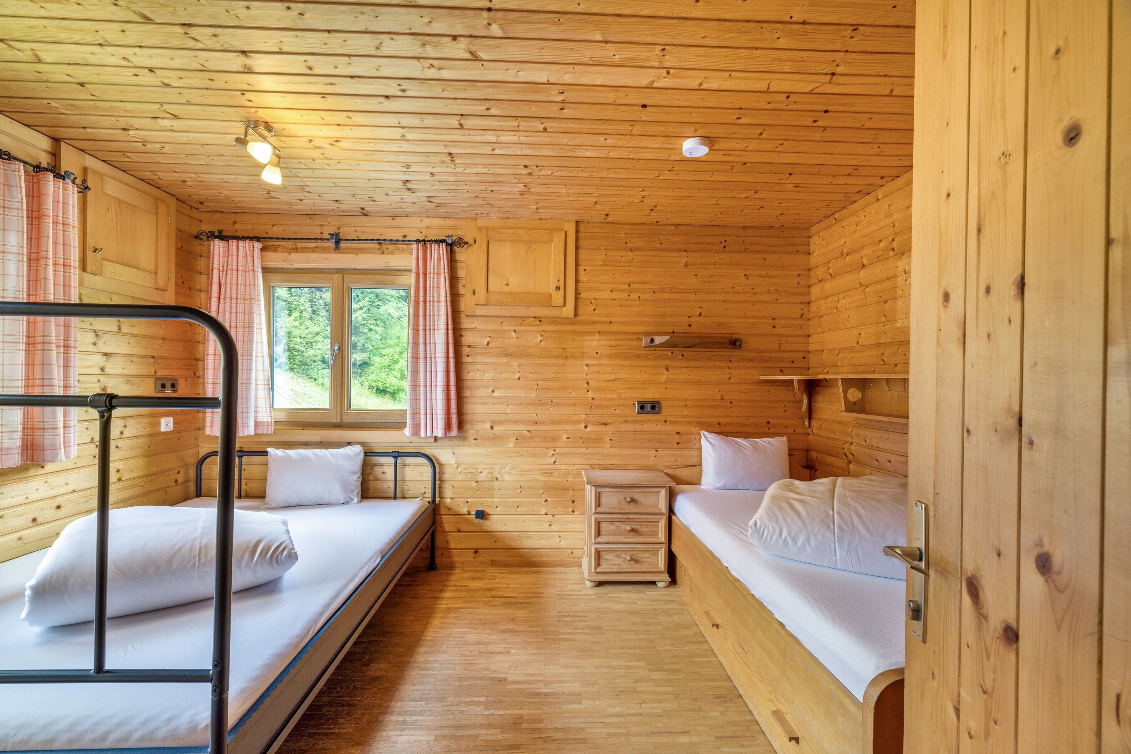 A wooden cabin room with bunk beds, a small dresser, and a window with curtains.