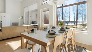 Een moderne keuken met een eettafel voor vier personen, grote ramen met uitzicht op bomen en een waterpartij, en een gezellige zithoek met een bank en een lamp.