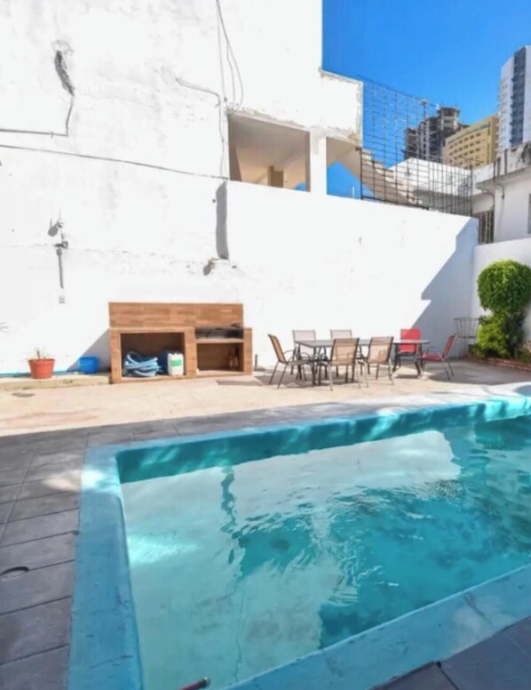 Ein Swimmingpool mit Holzterrasse, Gartenmöbeln und einer weißen Wand mit Fenster.
