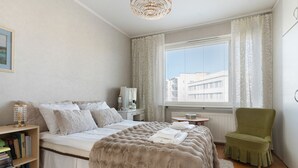 A bedroom with a bed, a green armchair, a bedside table with books, a window with blinds, and a view of buildings outside. - Aurora City Apartment 3bdr By Lapland Host (Rovaniemi)
