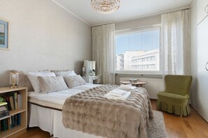 Ein Schlafzimmer mit einem Bett, einem grünen Sessel, einem Nachttisch mit Büchern, einem Fenster mit Jalousien und Blick auf Gebäude.