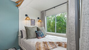 A bedroom with a bed, two wall-mounted lamps, a window with curtains, and a view of trees outside.