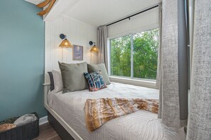 Ein Schlafzimmer mit einem Bett, zwei Wandlampen, einem Fenster mit Vorhängen und Blick auf Bäume.
