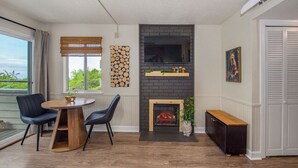 A modern living room with a fireplace, a round wooden table, two chairs, a TV mounted on a brick wall, and a window with a view.