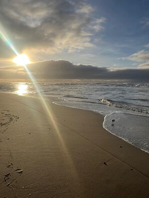 Uma praia com luz do sol refletindo na água, céu claro e pegadas na areia.