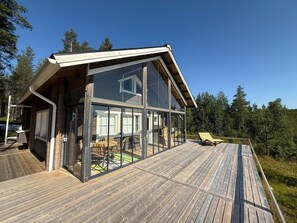 A modern house with large glass windows, a wooden deck, and a view of the surrounding forest.