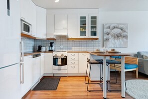 Una cocina compacta con armarios blancos, microondas, estufa y cafetera. Hay un comedor con mesa y sillas, y un sofá gris al fondo.