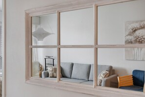 Una habitación con una ventana de seis divisiones, un sofá gris con cojines, una silla de madera, una lámpara colgante y un estante fijado a la pared.
