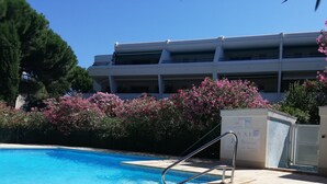 Uma piscina com uma escada de metal, rodeada por arbustos floridos cor-de-rosa, e um edifício alto ao fundo.