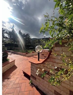Ein Ziegelweg führt zu einem Gartenbereich mit einem kugelförmigen Objekt an einer Wand, umgeben von viel Grün.