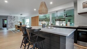 A modern kitchen with a large island, bar stools, pendant lighting, and a view into the dining area. - Designer Luxury 3BR with Pool and Hot Tub, Walk to Main St (Fredericksburg)