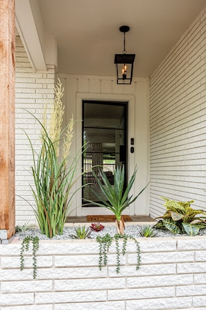 A white brick exterior with a black door, a hanging lantern, and various plants. - Designer Luxury 3BR with Pool and Hot Tub, Walk to Main St (Fredericksburg)