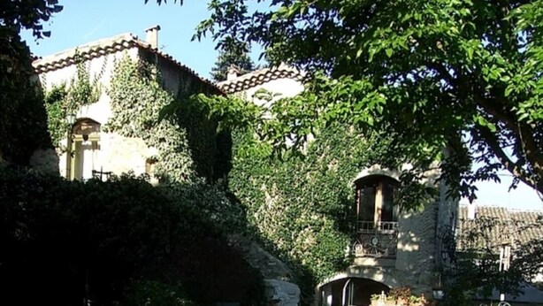 Un espacio de comedor al aire libre con sillas y mesas de metal, rodeado de vegetación y un edificio de piedra al fondo.