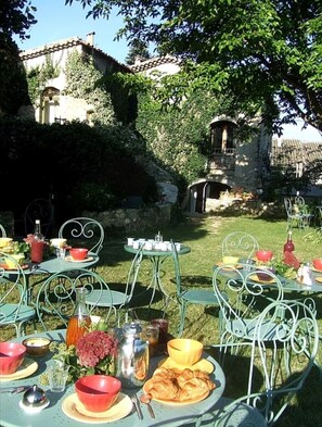 An outdoor dining area with metal chairs and tables, surrounded by greenery and a stone building in the background.