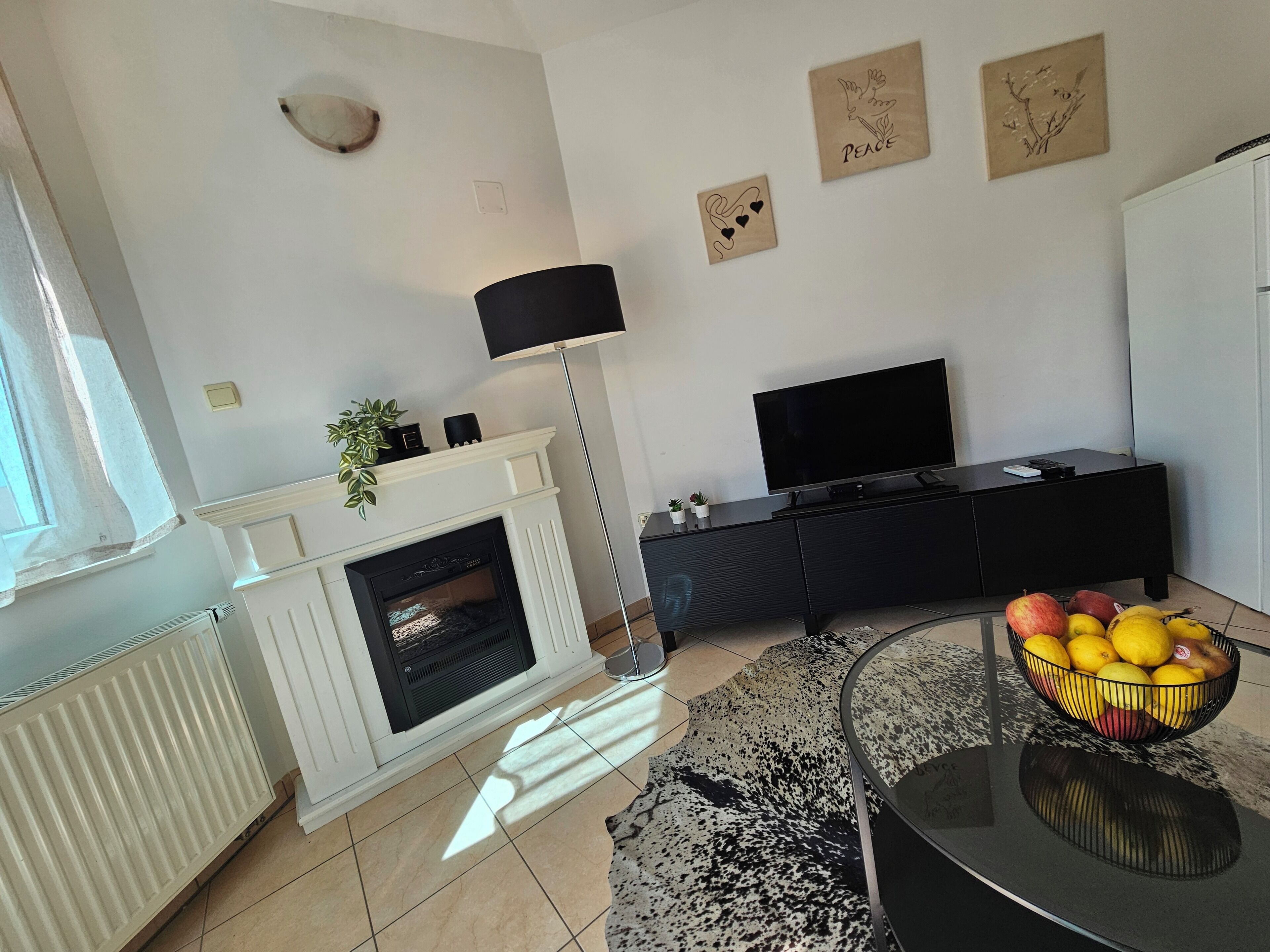 A modern living room with a fireplace, a flat-screen TV, a black entertainment unit, and a fruit bowl on a glass table.