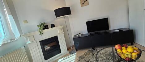 A modern living room with a fireplace, a flat-screen TV, a black entertainment unit, and a fruit bowl on a glass table.