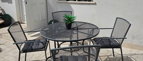 Une terrasse avec une table ronde en métal et quatre chaises, une plante en pot posée sur la table, et une fenêtre dont l’encadrement est orné de plantes.