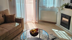 Un salon avec un canapé, une table basse en verre sur laquelle on a posé des fruits, une cheminée et une vue par la fenêtre.