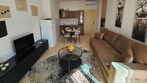 Un salon comprenant un canapé, une table basse avec des fruits, un téléviseur et un coin cuisine avec une table à manger et des chaises.