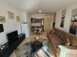 A living room with a sofa, a coffee table with fruit, a TV, and a kitchen area with a dining table and chairs. - Luna Mare, One-Bedroom Apartment on Čiovo (Trogir)