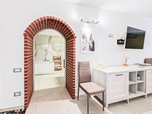 Un bagno con parete in marmo, una vasca da bagno bianca, uno scaffale in legno, una sedia, un mobile vanitario con specchio e un televisore montato a parete.