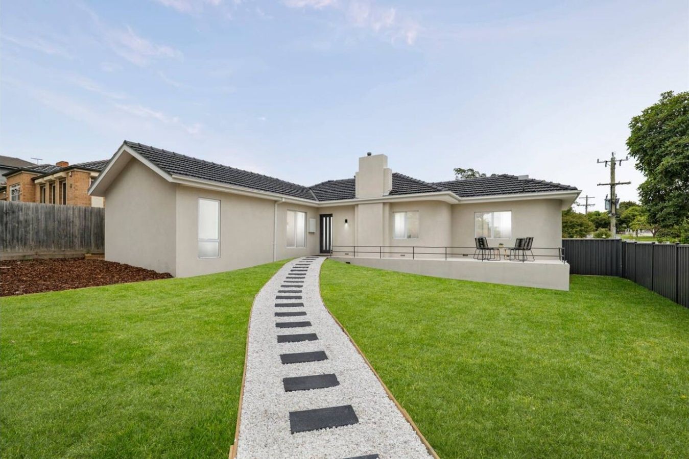 Charming single-level home with a lush front lawn and a stepping-stone path leading to the entrance. A raised front patio with outdoor seating offers a peaceful spot to enjoy morning coffee or a quiet evening outside.