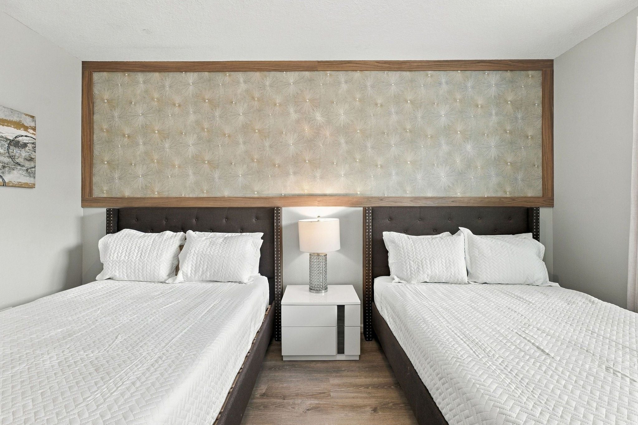 A hotel room with two beds, a nightstand with a lamp, and a large artwork above the beds.