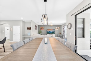 A modern dining room with a long wooden table, grey upholstered chairs, a central light fixture, and a cityscape painting on the wall. - Sol117-5484-6-pg - Solterra's Dream Home (Davenport)