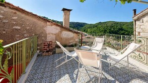 Una terraza con suelo de baldosas, sillas blancas, pared de ladrillo y vistas a colinas verdes.