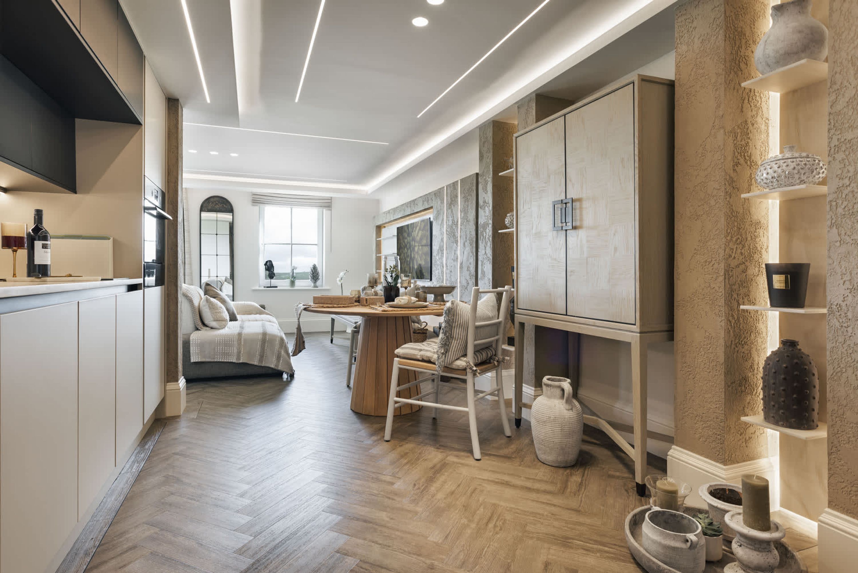 A modern kitchen with wooden flooring, a dining area with a table and chairs, and a living space with a sofa and a large mirror.