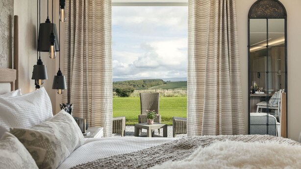 A bedroom with a large bed, a view of the outdoors, and a dining area visible through the window. - The Hide Ridgestone (Penrith)