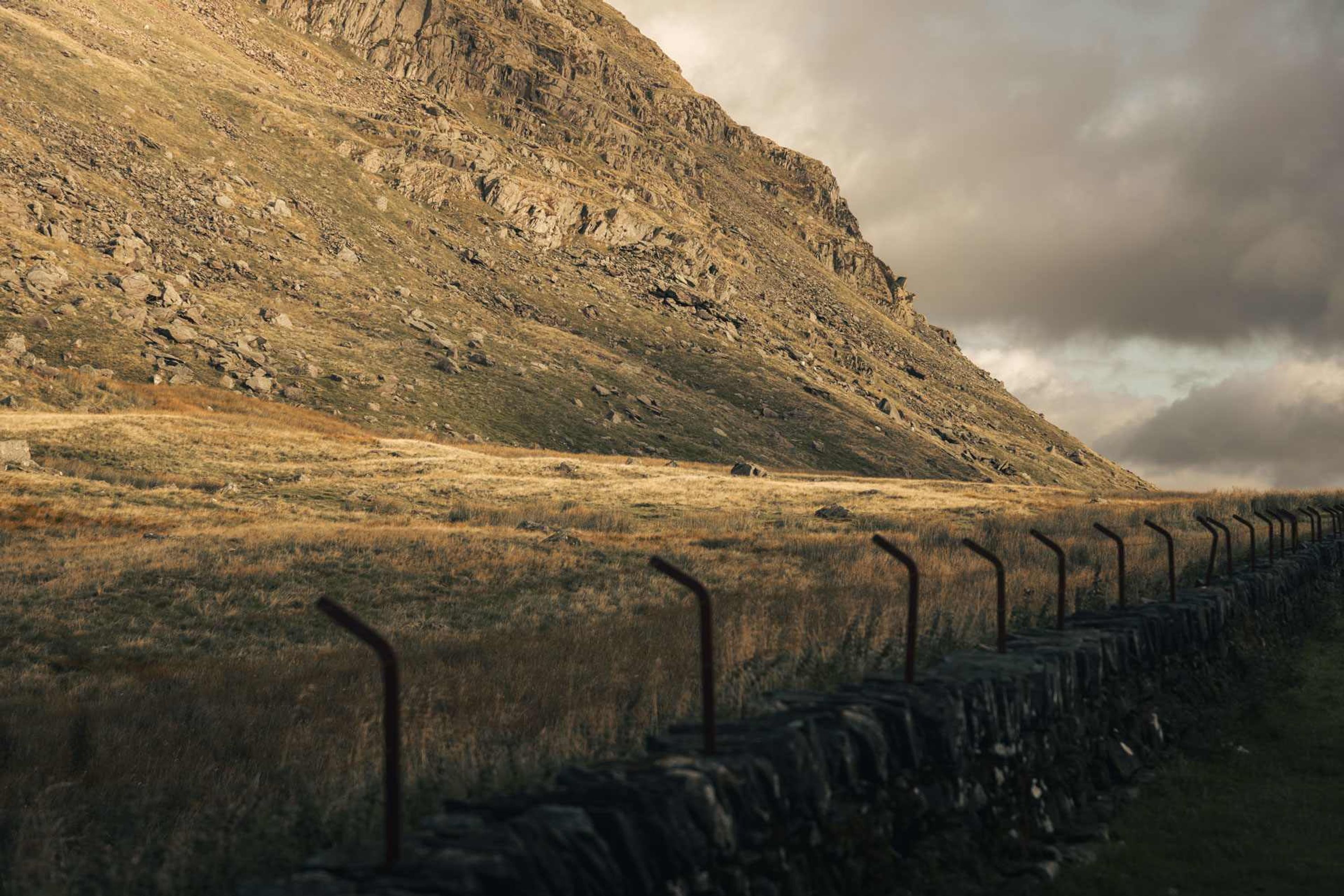 A stone wall with metal railings, a grassy field, and a rocky hillside.