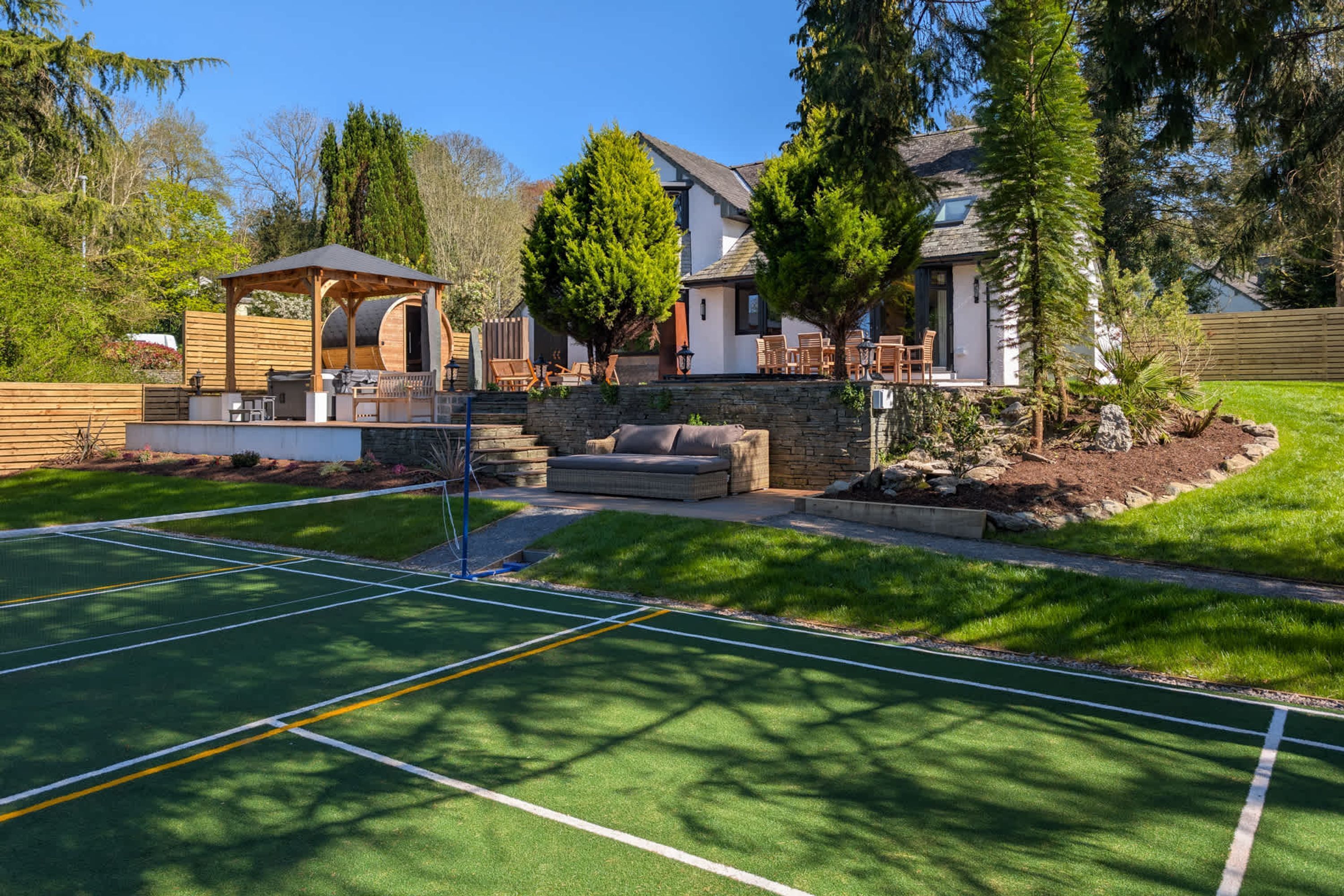 A tennis court with a wooden pavilion, a stone wall, and a house with a sloped roof.