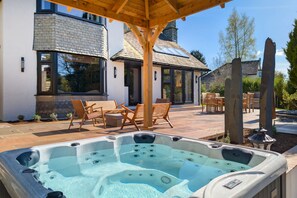 A hot tub in a covered outdoor area with a patio, chairs, and a building with large windows. - Wyke View Ridgestone (Windermere)