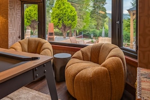 Una habitación con mesa de billar, dos sillones tapizados, una pequeña mesa redonda y vistas a un jardín con árboles y un pabellón.